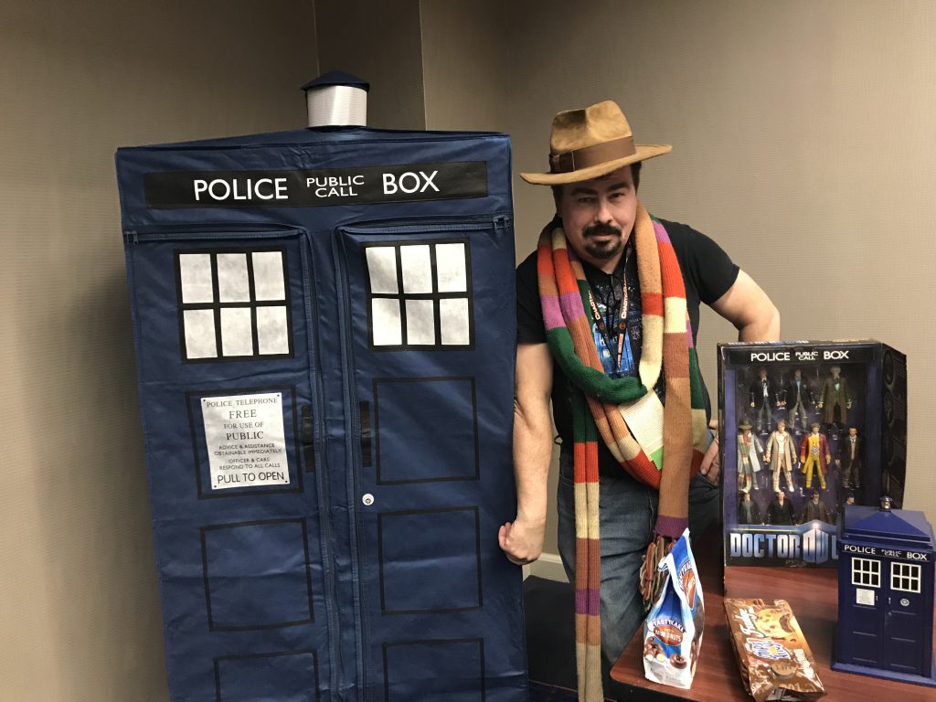 Doctor Who fan Roger Riddle dressed in various character elements, standing next to a collection of toys and memorabilia from the television programme.